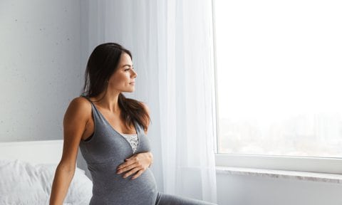 Mujer embarazada en el primer trimestre sentada en la cama mirando por la ventana