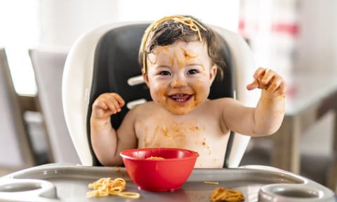 Bebé sentado en la trona de la cocina comiendo un plato de espaguetis.