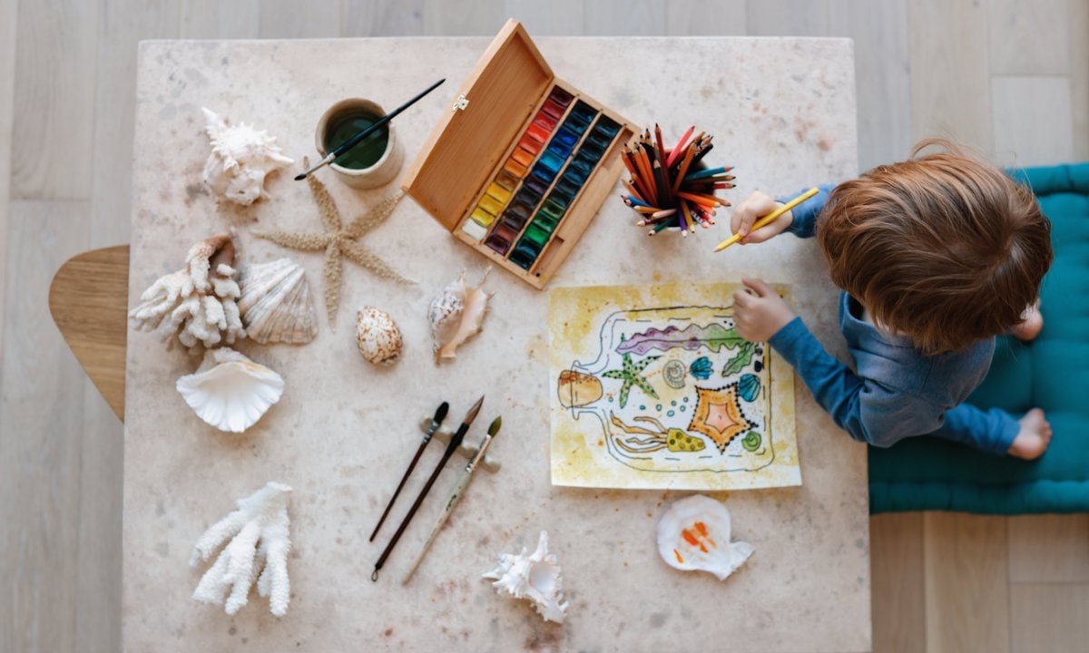 Niño en una mesa haciendo manualidades con pinturas y conchas
