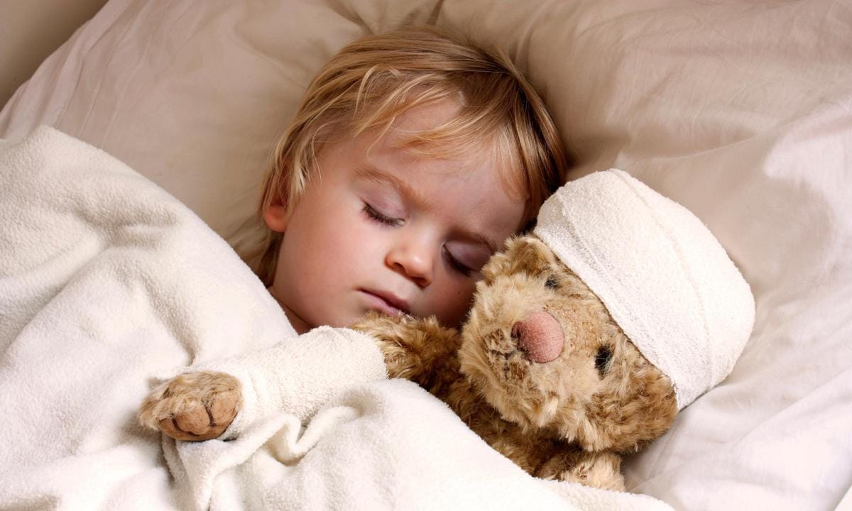 Niño enfermo en la cama al lado de un oso con la cabeza vendada