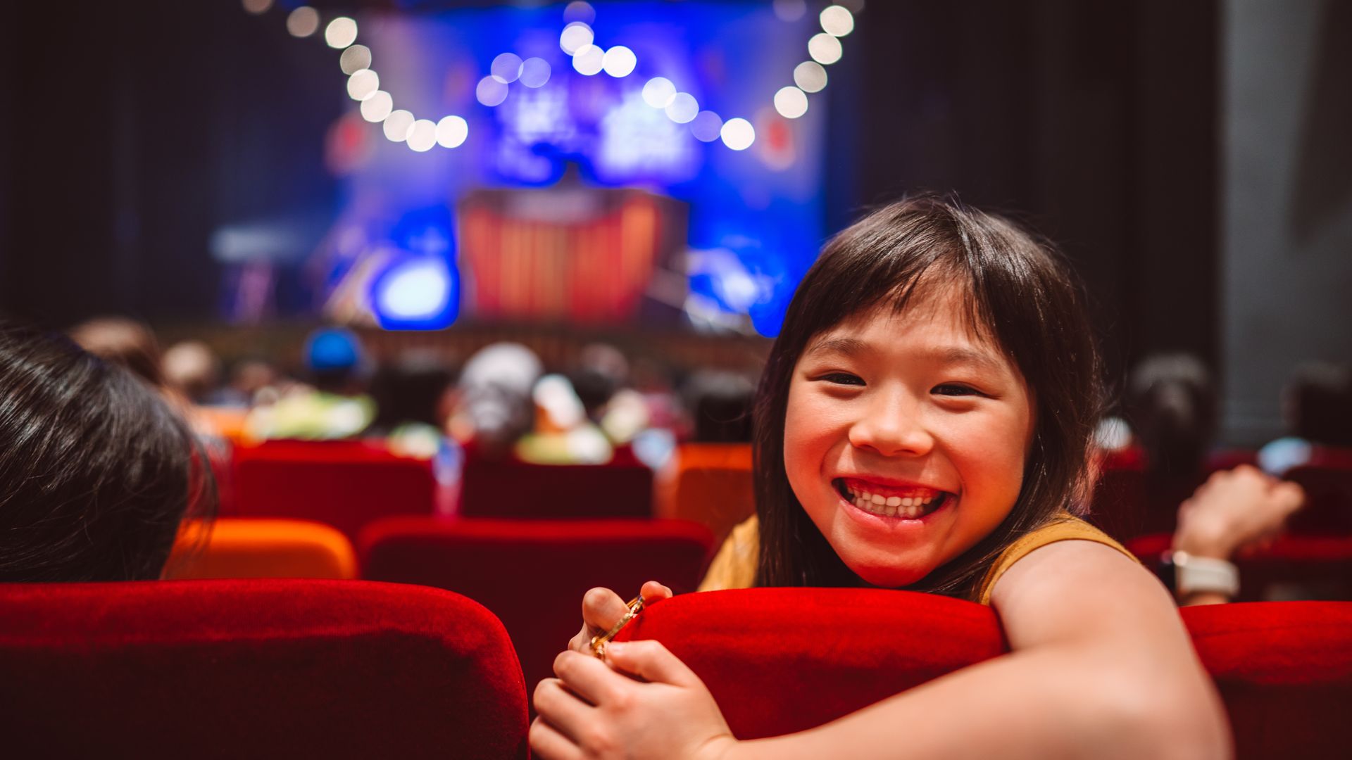 Cheerful young girl beams with excitement while seated in a red velvet theatre chair, eagerly awaiting a live performance in a vibrant stage setting. The photo captures the wonder and anticipation of childhood, highlighting themes of arts education, cultural enrichment, and family entertainment. 