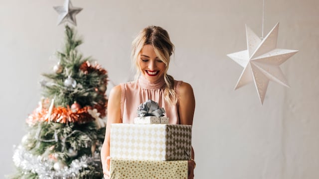 Cheerful smiling female standing next to the decorated Christmas tree and holding wrapped gift boxes in hand