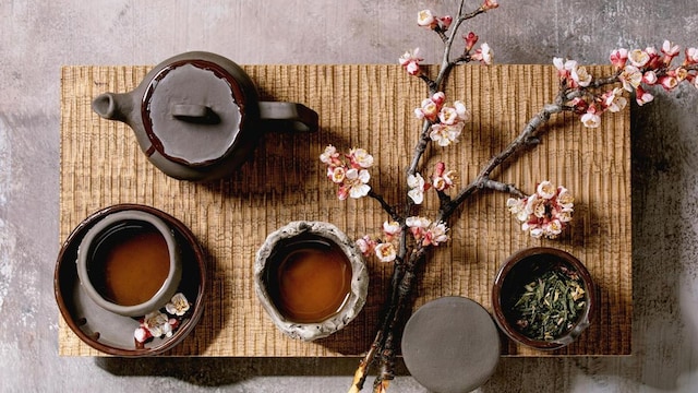 Tea drinking wabi sabi japanese style dark clay cups and teapot on wooden tea table with blooming cherry branches. Grey texture concrete background. Flat lay. space