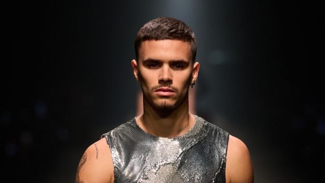 Romeo Beckham walking down the runway wearing a metallic mesh sleeveless top, black trousers, studded black gloves, and displaying his tattooed arms.