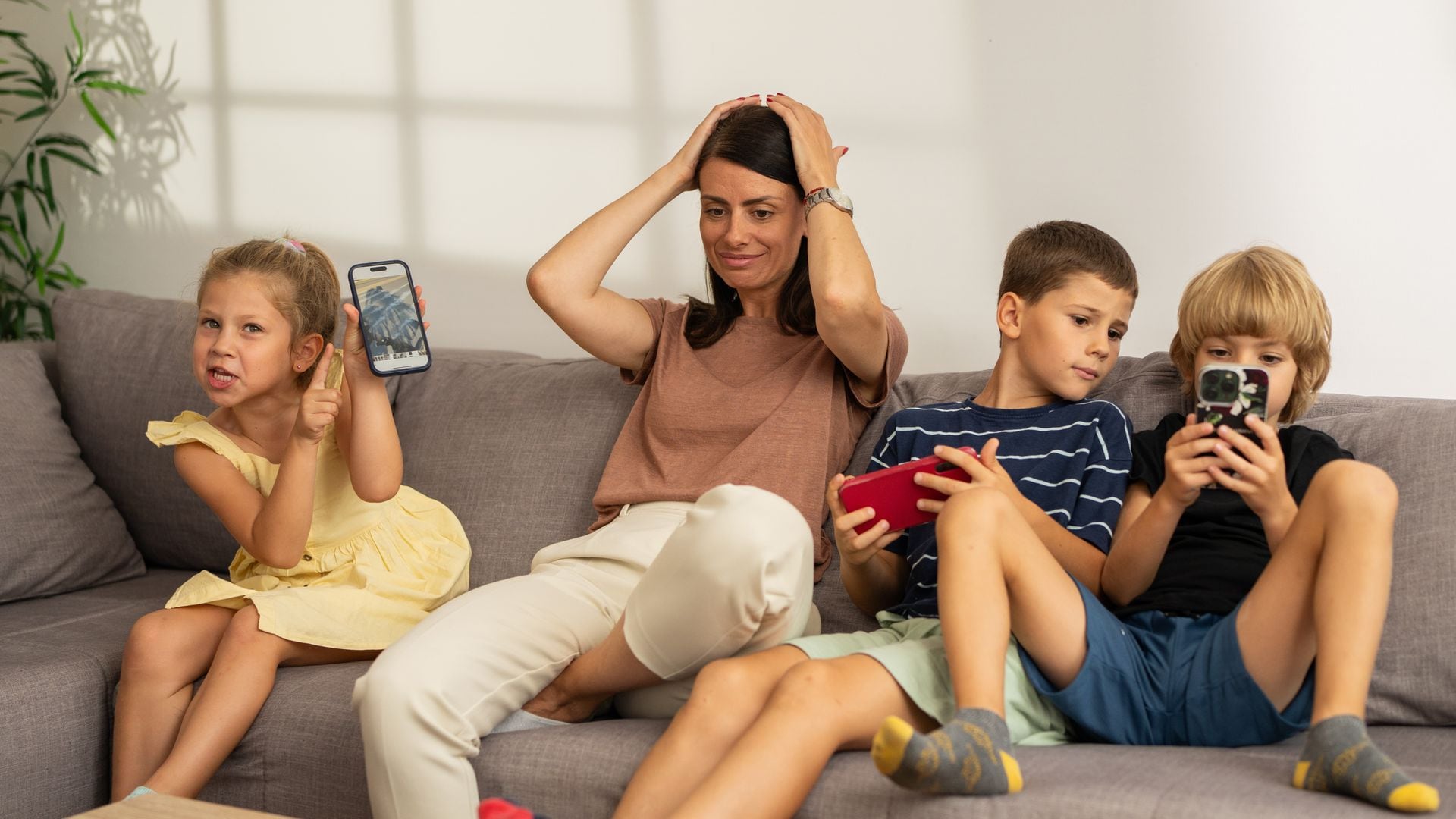 Tired young mother sits on couch and holds her head while her three kids play games on smartphones 