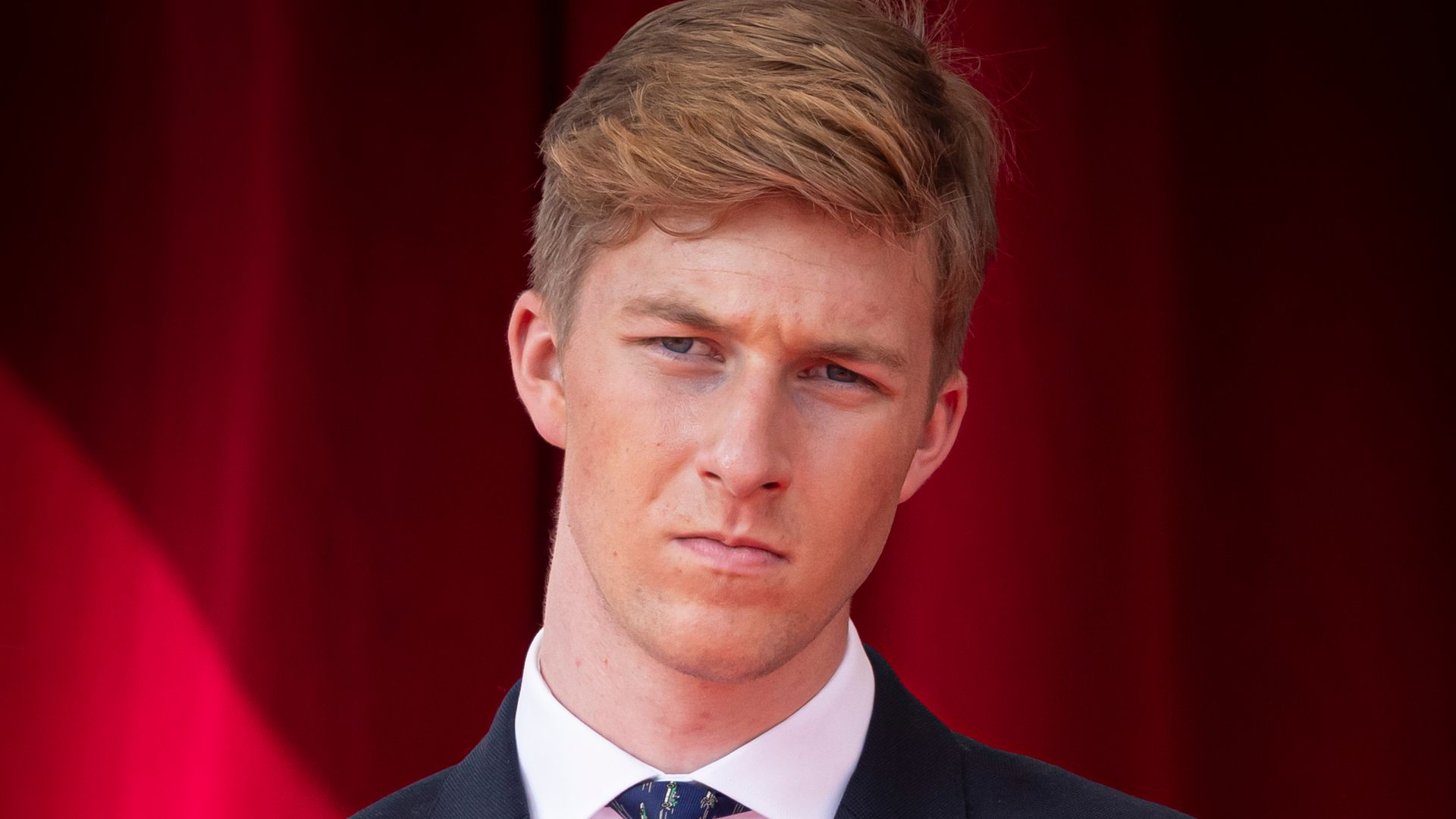 Prince Emmanuel of Belgium in a dark suit and striped tie, standing against a red curtain backdrop.