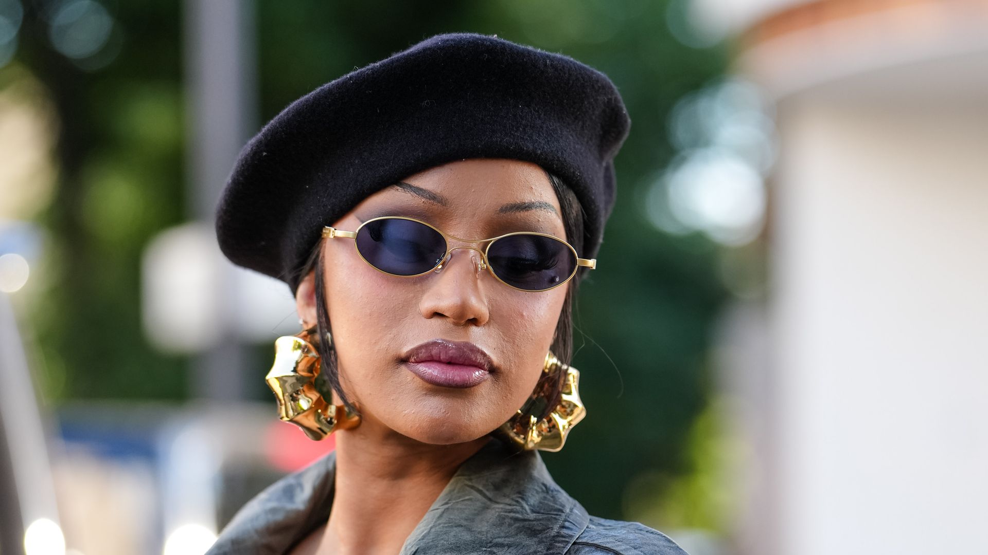 Cardi B wears a black beret and small, oval sunglasses with tinted lenses. Large, gold hoop earrings are visible. The outfit features a long, olive green trench coat with a double-breasted design, buttoned closure, and a matching belt at the waist. 