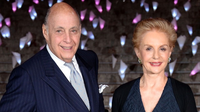 Reinaldo Herrera and Carolina Herrera pose together at an elegant event, both dressed in formal attire, smiling against a sophisticated backdrop.