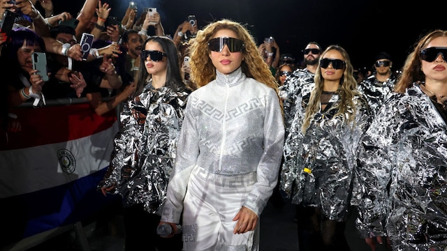Shakira walks through the crowd in a silver outfit and sunglasses, surrounded by dancers in metallic attire, as fans cheer during her Las Mujeres Ya No Lloran tour.