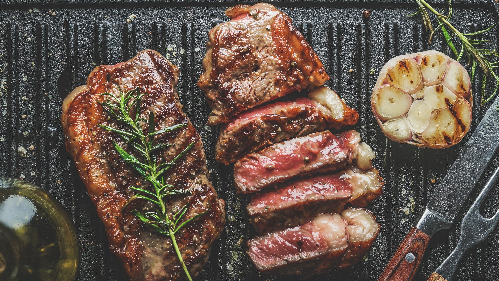 Close up of sliced roasted medium rare barbecue steak with rosemary, roasted garlic and cutlery on rustic iron grill. Cooking at home with fresh meat and flavorful herbs. Top view. 