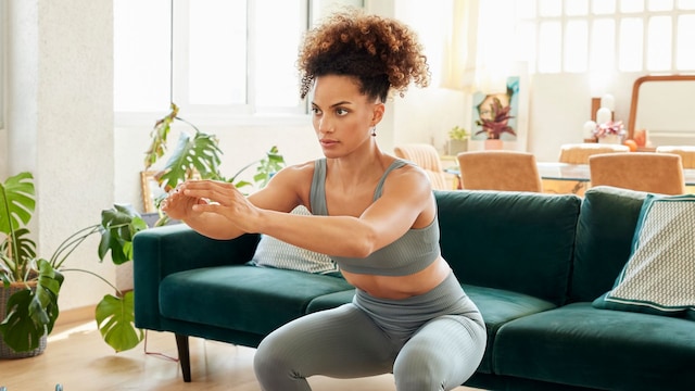Full length of fit female doing squats in front of sofa. Young woman in sportswear is exercising at home. She is in living room. 