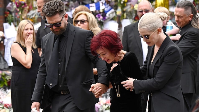Sharon Osbourne, devastated by grief, is held up by her children Jack and Kelly as the family says a painful goodbye. Surrounded by mourners and a flood of flowers, their sorrow is clear. This is a final farewell frozen in a quiet, powerful moment.