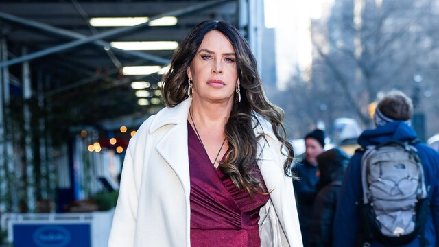 Karla Sofía Gascón walking in a city street wearing a burgundy satin dress and a white coat, carrying a black handbag with a confident expression