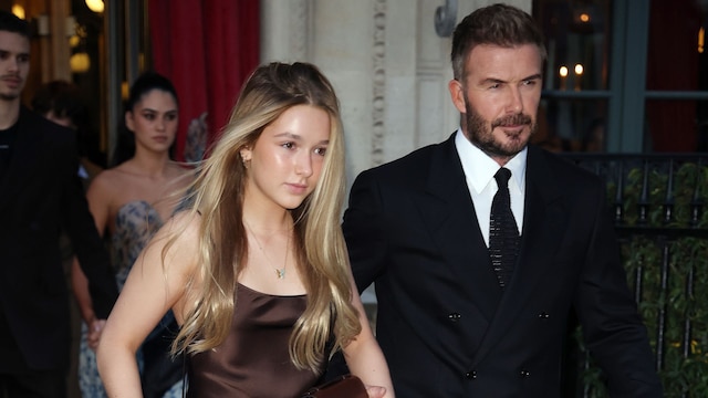 Harper Beckham in a brown satin dress walking beside her father, David Beckham, who is dressed in a black suit, at Paris Fashion Week 2024.