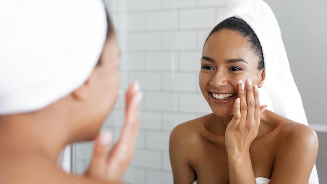 Happy woman putting on facial moisturizer with hand in bathroom mirror