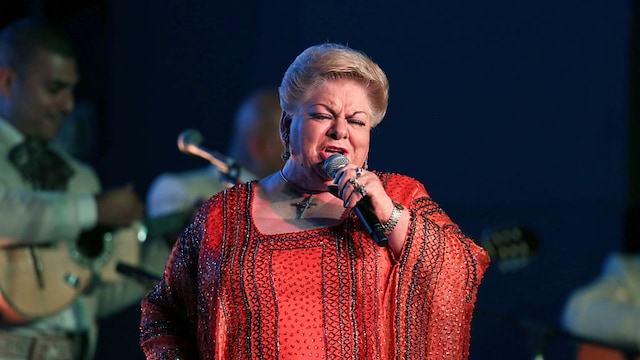 Paquita la del Barrio singing on stage in a red embroidered outfit, with mariachi musicians playing in the background.