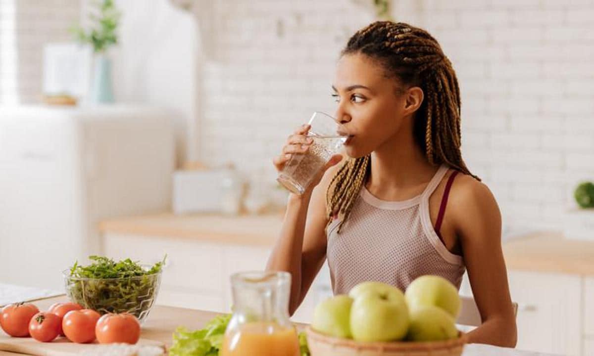 Joven bebe agua en su cocina en medio de alimentos sanos