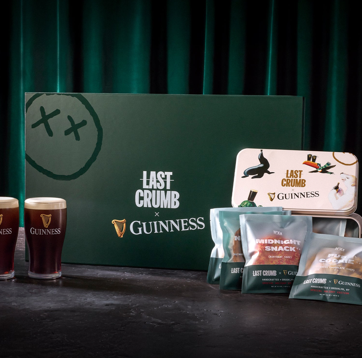 Two pints of Guinness, a green box labeled 'Last Crumb x Guinness', and several snack packages on a dark surface.