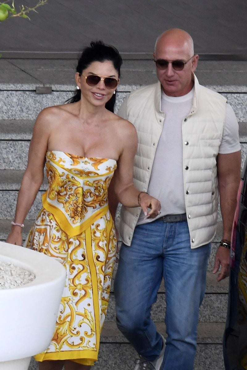 A BIT WINDY! Lauren Sanchez and Amazon founder/billionaire partner Jeff Bezos arrive at the Cagliari Airport to attend the Dolce and Gabbana show. The two showed a bit of PDA as Jeff assisted Lauren to the car and got an eye full of Sanchez's... assets. Lauren is wearing a Dolce & Gabbana dress.