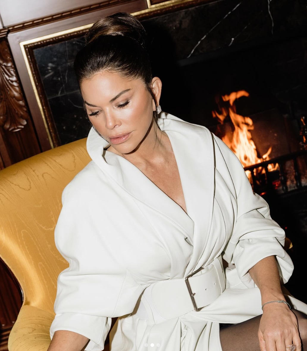 The images in the post show a confidently glamorous Lauren Sánchez Bezos wrapped in a sculptural white coat, lounging by a crackling fireplace.
