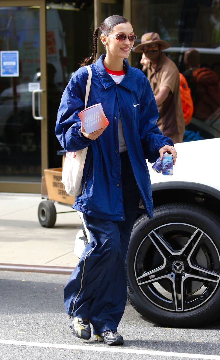 Bella Hadid wearing a blue oversized outfit with a white and red tee. Bella completes her look with sunglasses and braided hair.