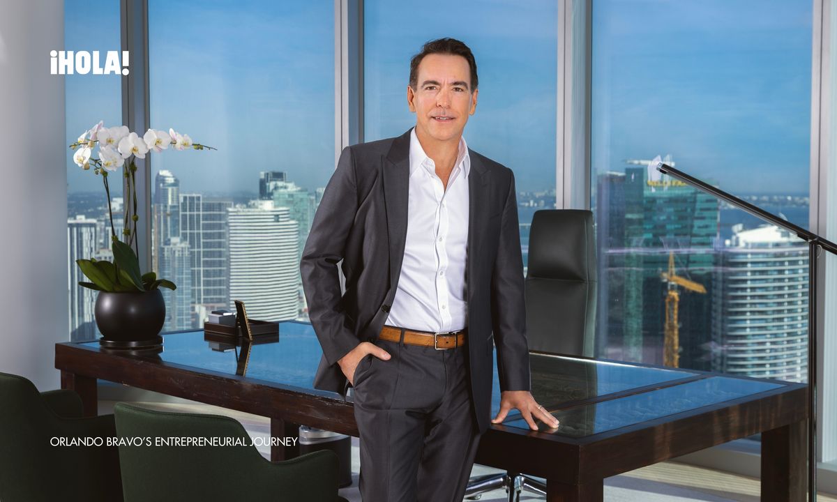 Orlando Bravo, dressed in a dark suit, leans against his desk in a high-rise office with a panoramic view of Miami’s skyline in the background.