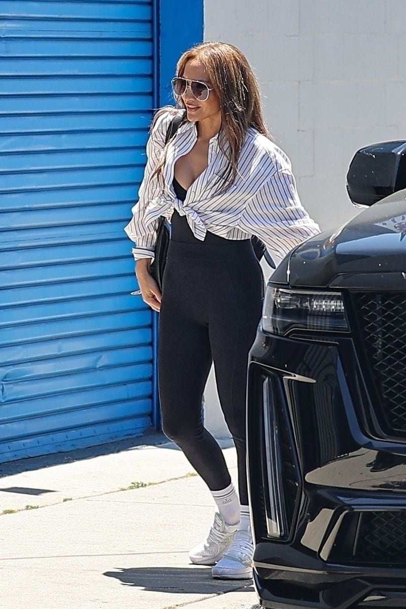 JLo was pictured arriving at a studio this afternoon, wearing a striped dress shirt, black leggings, and white designer sneakers.