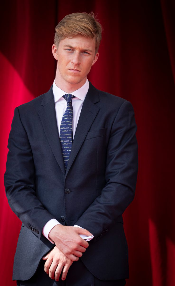 Prince Emmanuel of Belgium in a dark suit and striped tie, standing against a red curtain backdrop.