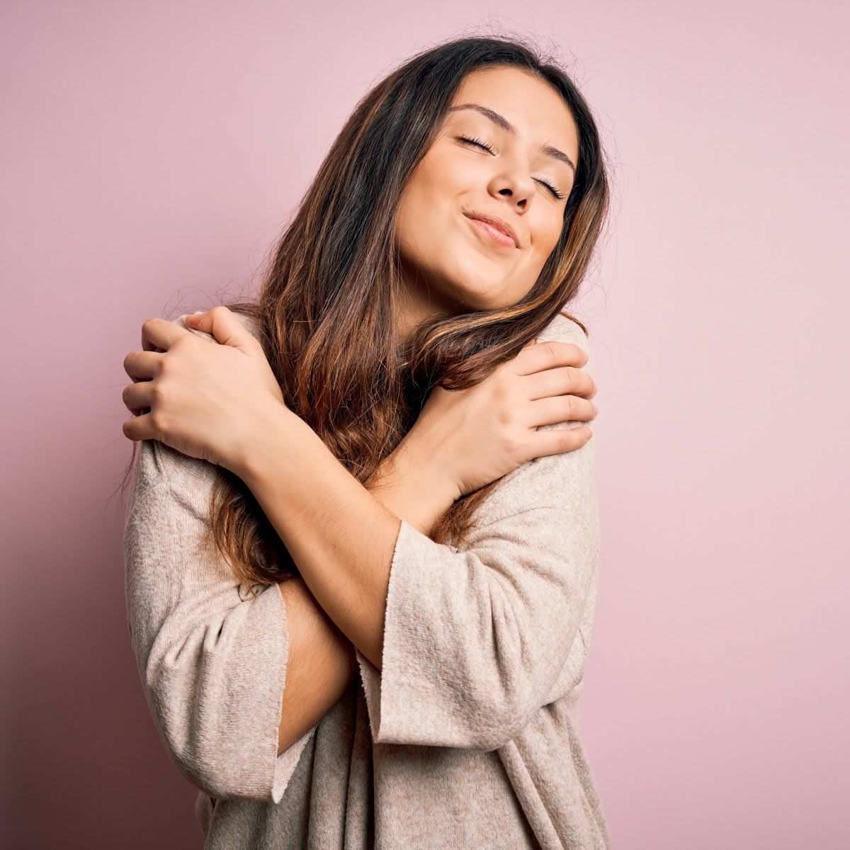 Young beautiful brunette woman wearing casual sweater standing over pink background Hugging oneself happy and positive, smiling confident. Self love and self care