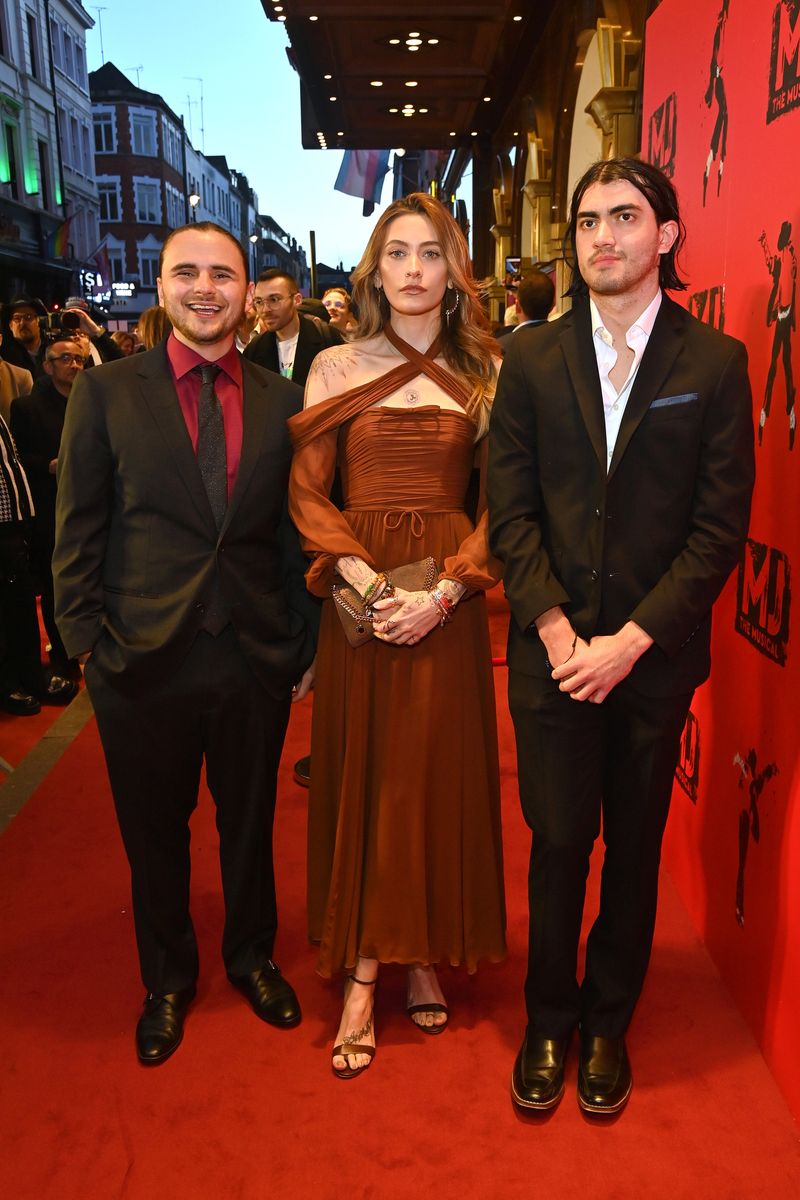 Three people on a red carpet: Paris Jackson in a brown dress, flanked by two men in suits.