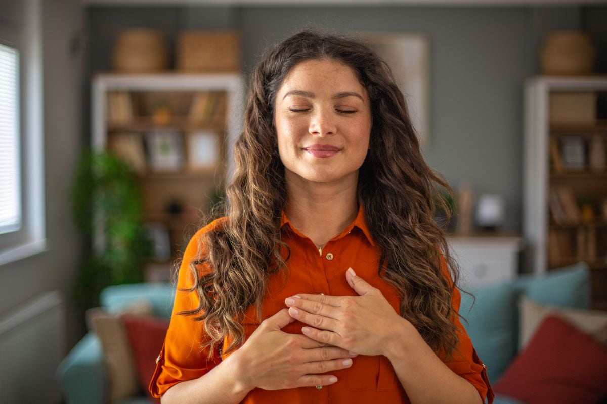 Young woman practicing mindfulness at home, holding hands on chest and enjoying peace