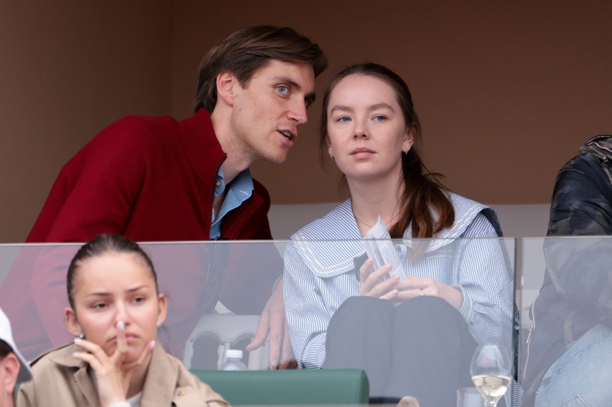 Ben-Sylvester Strautmann in a red sweater leans toward Alexandra de Hannover, who wears a blue-and-white striped top and holds a program, as the two sit in a stadium-style seating area with a glass of wine on the ledge in front of them.