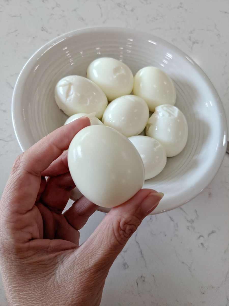 Woman's hand holding a peeled Hard-boiled egg in front of bowl of eggs