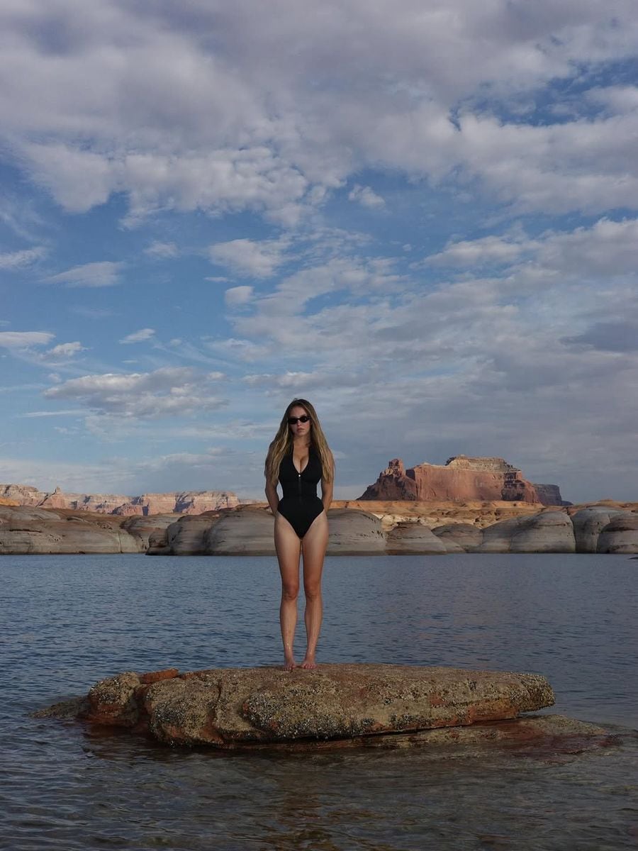 Like a desert goddess on water, Sydney Sweeney strikes a powerful pose in a sleek black one-piece while standing on a lone rock in the middle of the lake.