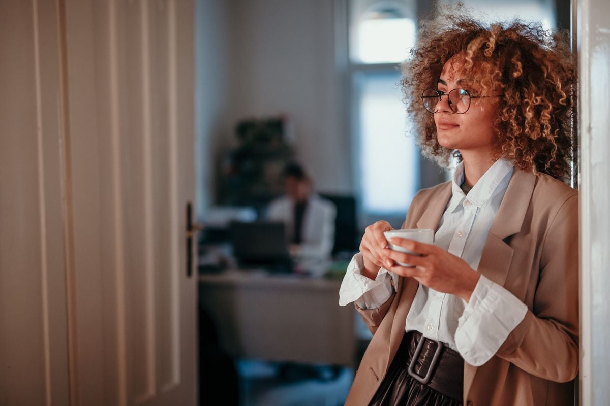 A waist up photo of a beautiful, cool-looking, fashionable dressed young adult business director leaning against the door, looking away with a coffee in her hands, contemplating