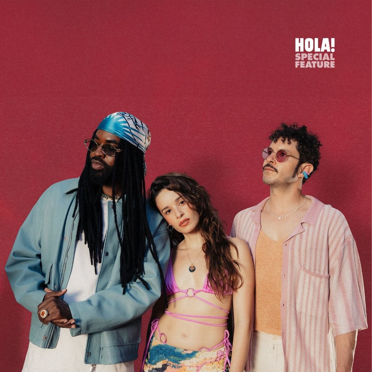 Three people stand together against a red background. The man on the left wears a blue jacket and a blue headwrap. The woman in the middle dons a pink bikini top and colorful skirt. The man on the right has curly hair and wears a pink striped shirt.