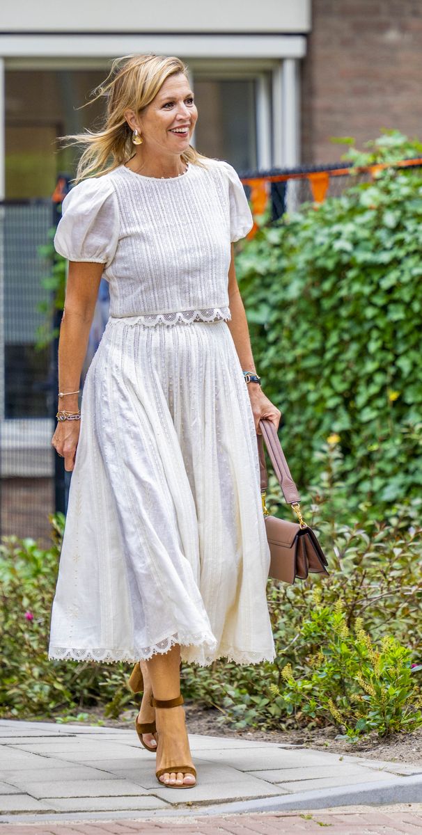 Queen Maxima looking elegant in a white dress paired with beige accessories, including a brown handbag and matching heels.