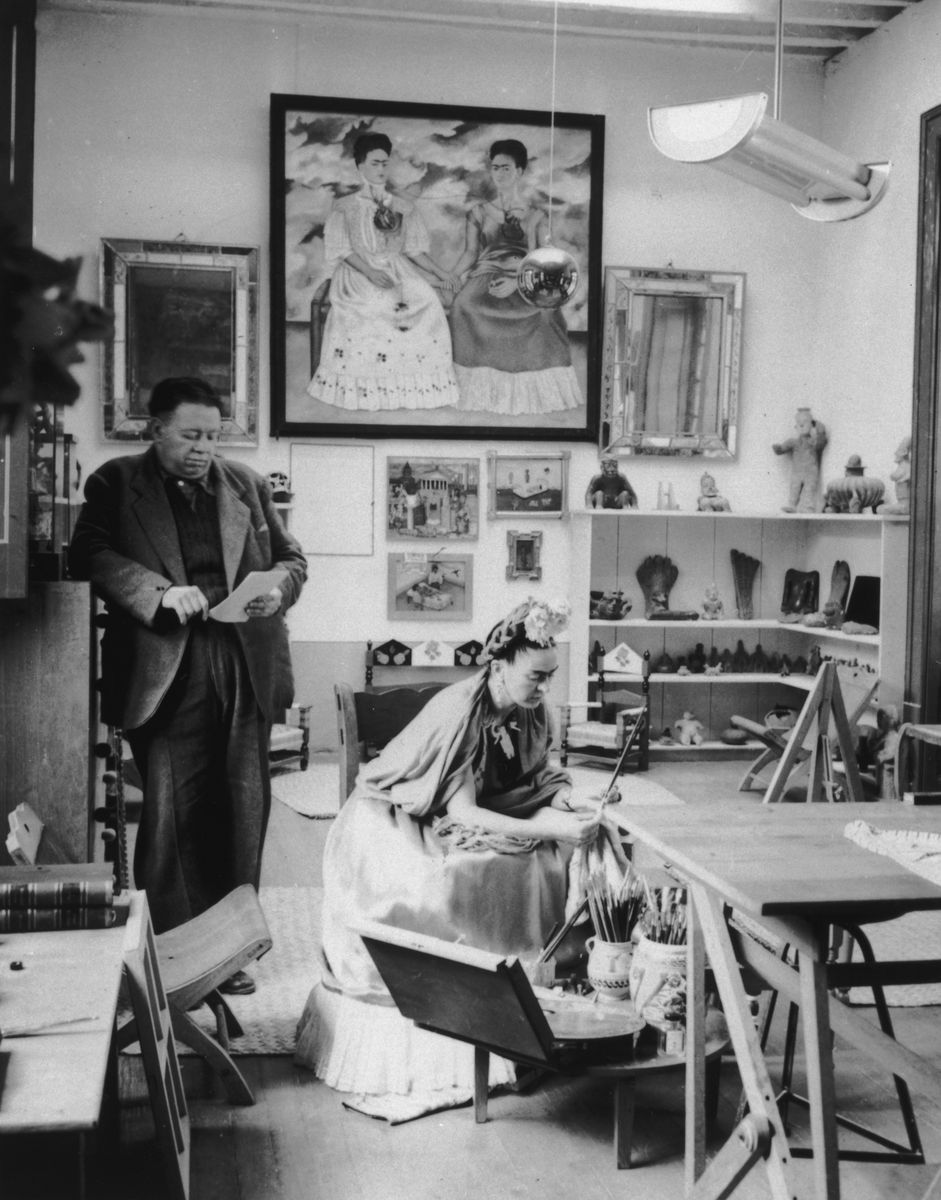 Married Mexican artists Diego Rivera and Frida Kahlo (1907 - 1954) read and work in a studio. Kahlo's self-portrait, 'The Two Fridas' (1939), hangs in the background with other works. 