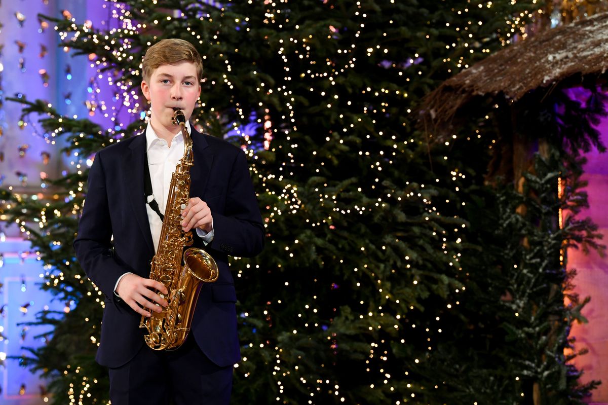 A young Prince Emmanuel of Belgium, in a navy suit, plays the saxophone in front of Christmas trees strung with lights