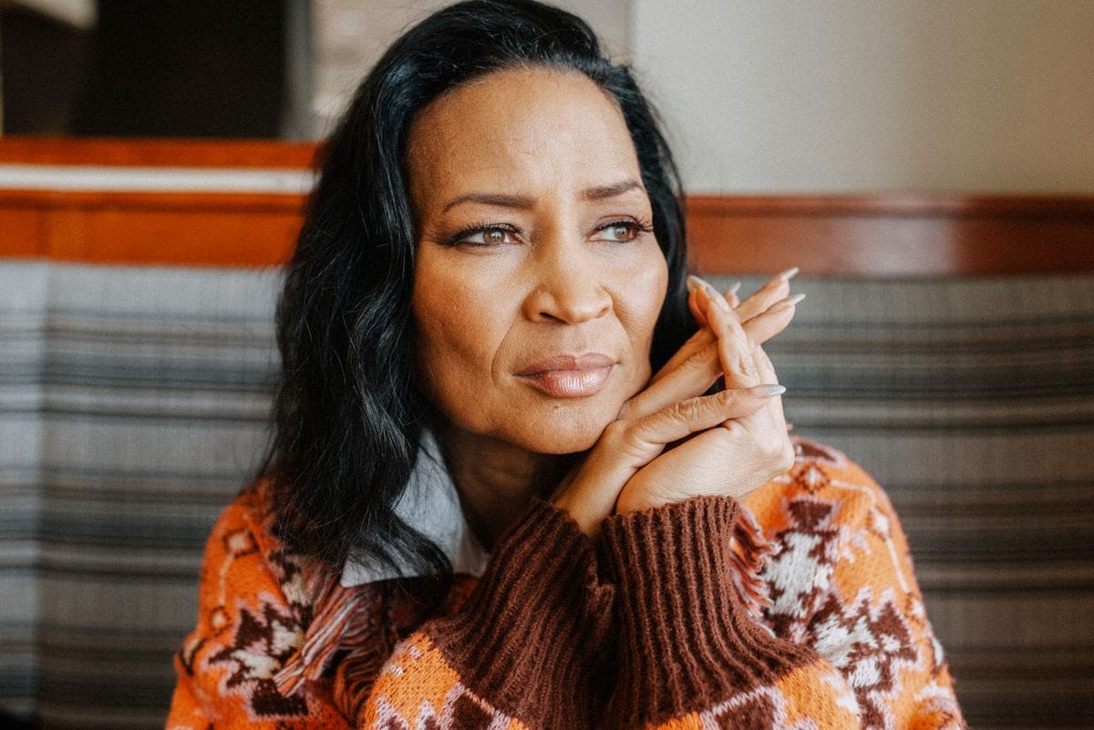 A woman gazes thoughtfully into the distance, her hands gently clasped under her chin. She wears a cozy, patterned sweater, adding warmth to the indoor setting.