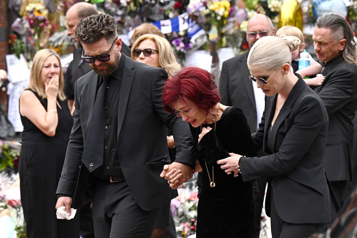 Sharon Osbourne, devastated by grief, is held up by her children Jack and Kelly as the family says a painful goodbye. Surrounded by mourners and a flood of flowers, their sorrow is clear. This is a final farewell frozen in a quiet, powerful moment.