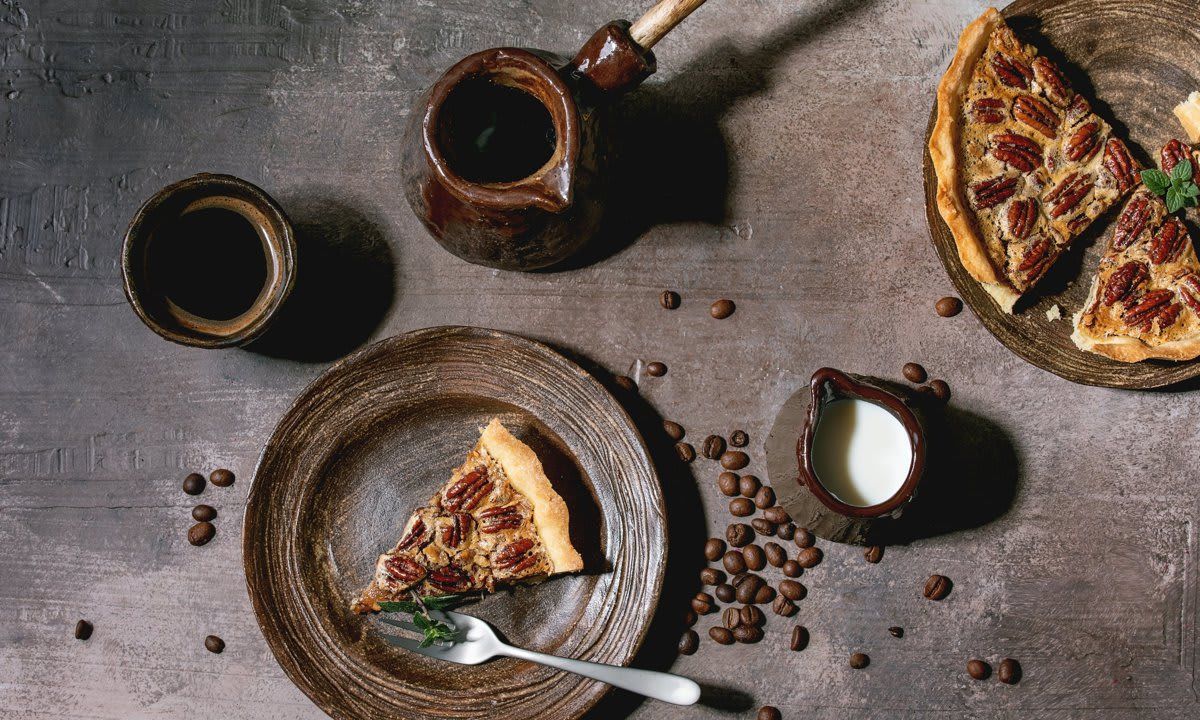 Piece of home baking classic american pecan caramel pie on ceramic plate, coffee cup, cezve, jug of cream over grey texture background. Flat lay, space