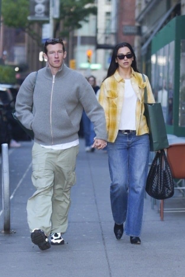 Dua is wearing a yellow plaid shirt over a white top, paired with blue jeans and sunglasses, while Callum wears a grey zip-up sweater with khaki cargo pants and colorful sneakers.