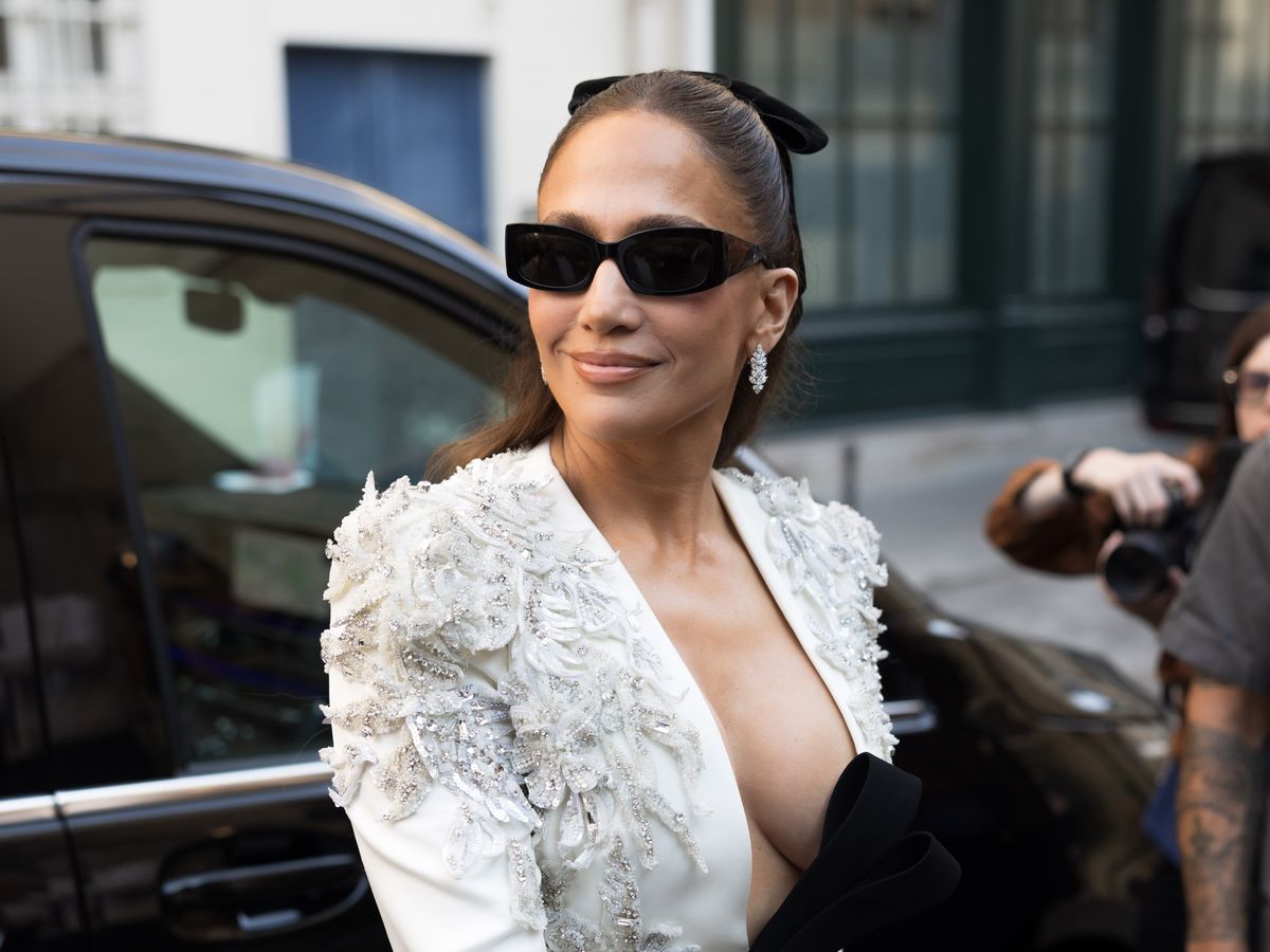 Jennifer Lopez stands by a car, wearing a white embellished top and black sunglasses.