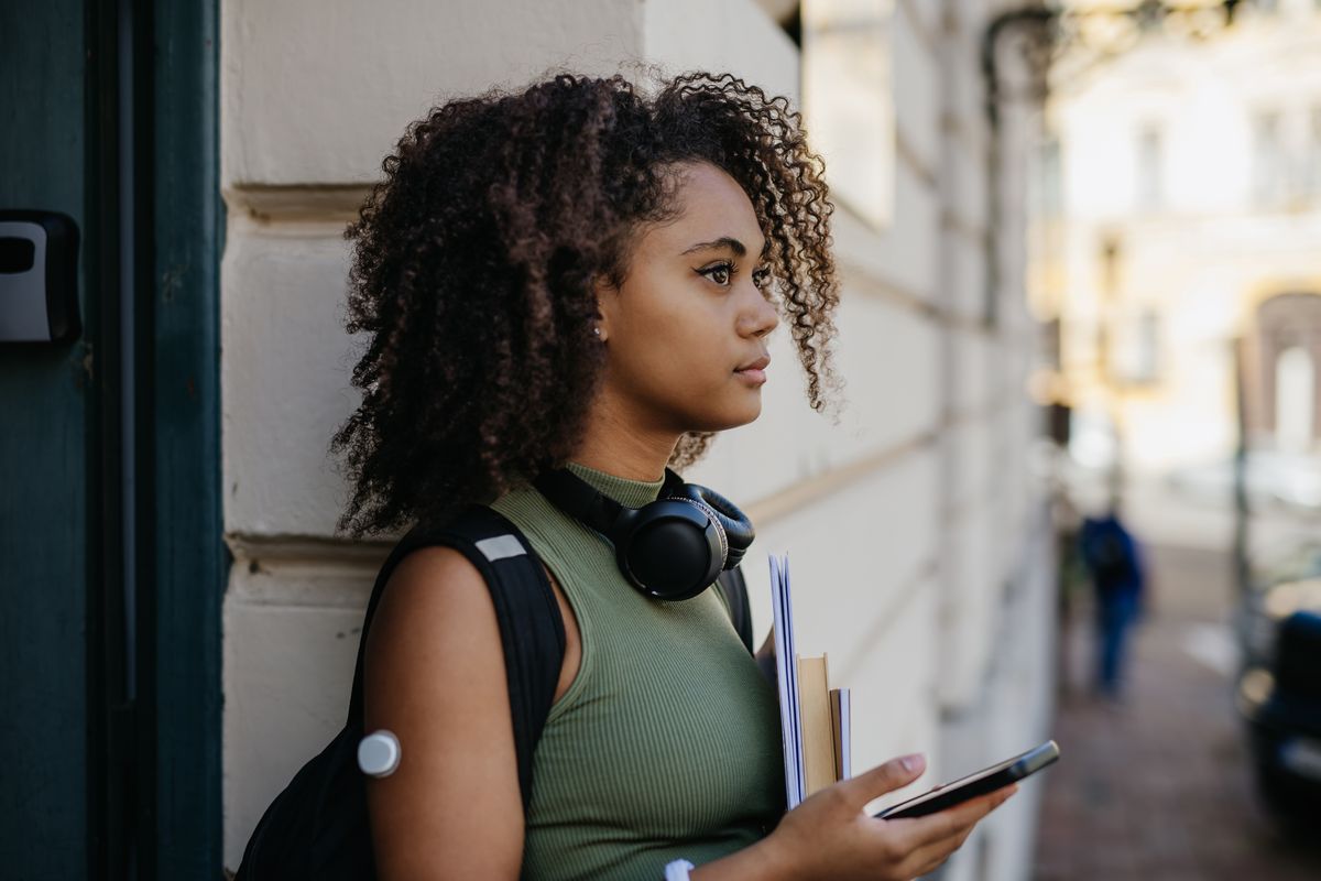 Portrait of teenage diabetic student standing outdoors in the city with smartphone in hand. Teenage girl wearing continuous glucose monitor sensor on her arm to keep her blood sugar level in check. Concept of teens and diabetes.