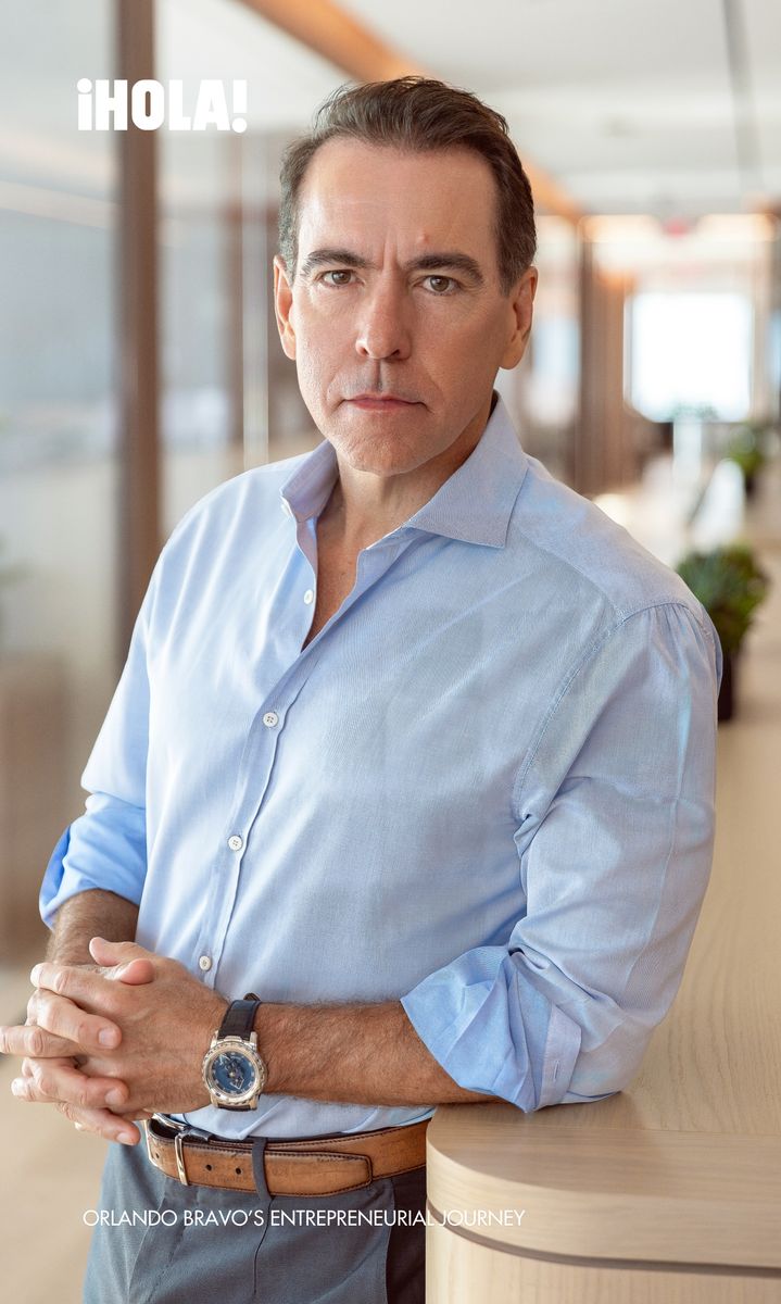 Orlando Bravo, dressed in a light blue shirt with sleeves rolled up, leans against a modern office desk with a determined expression, reflecting his entrepreneurial spirit.