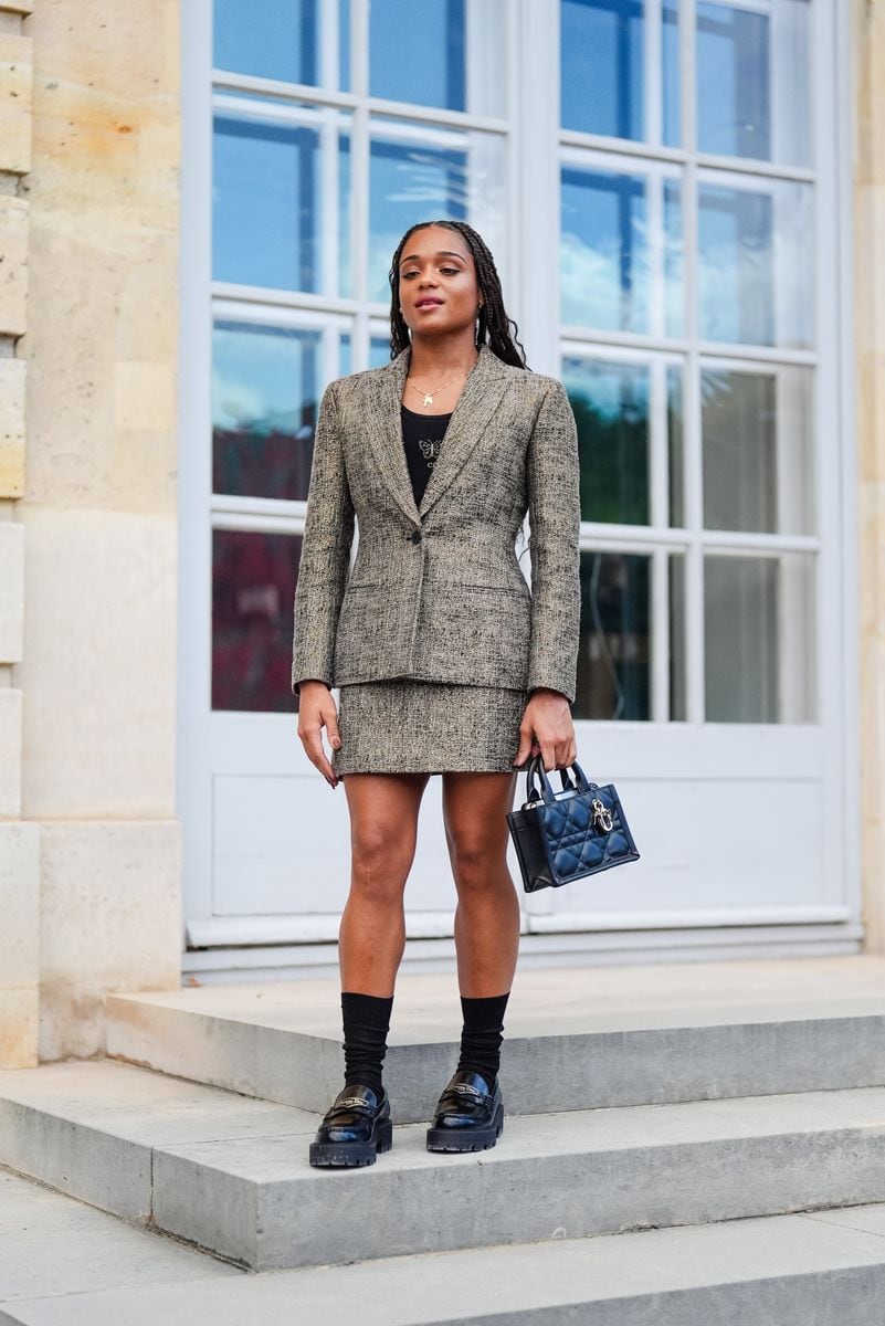 A guest wears gold necklace with cross pendant, black t-shirt with silver butterfly pattern, light brown pattern/print tailored blazer jacket, light brown pattern/print mini skirt, shiny black Dior Lady D-joy leather bag, black socks, shiny black Christian Dior loafer moccasin leather shoes, outside Dior, during the Paris Haute Couture Week Fall/Winter 2024-2025 on June 24, 2024 in Paris, France. (Photo by Edward Berthelot/Getty Images)