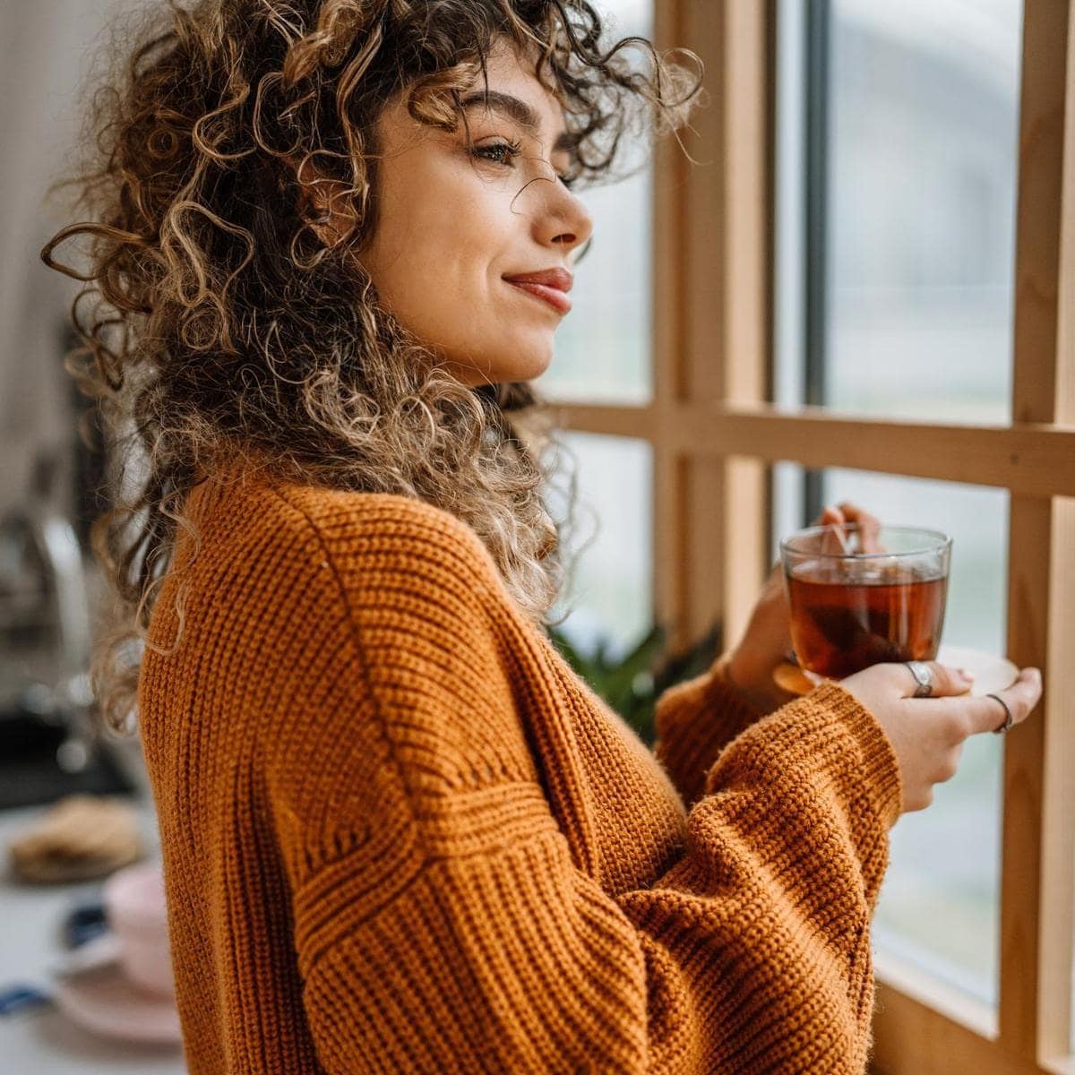 Young woman holding a cup of tea and standing by the window. Tea time rituals.