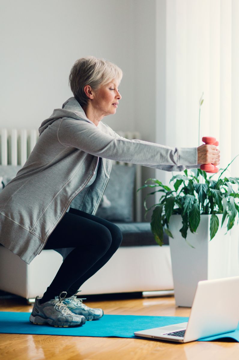 Mature woman exercising at home, learning exercises online with laptop. Doing squats with dumbbells.