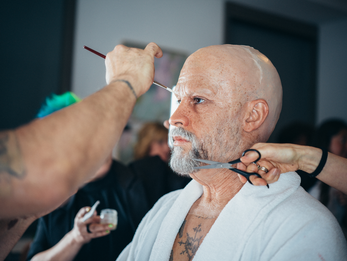 Man with bald head and beard getting makeup and hair styled by two people.